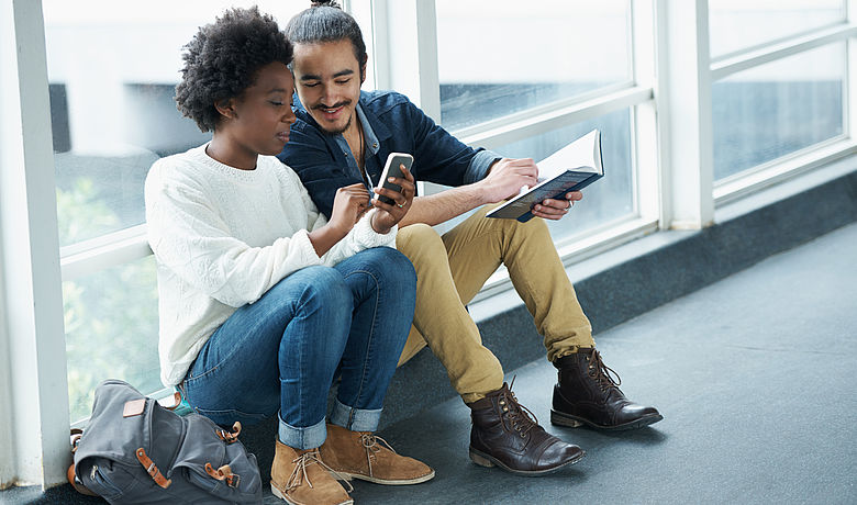 Zwei junge Menschen mit unterschiedlicher Hautfarbe sitzen zusammen und zeigen einander etwas auf einem Smartphone