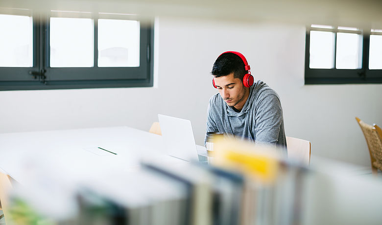 In einer Bibliothek arbeitet ein junger Mann mit Kopfhörern konzentriert an seinem Laptop 