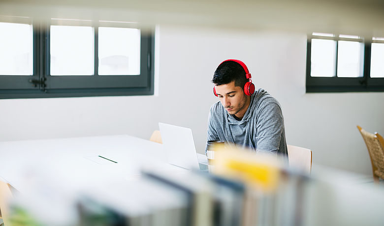 In einer Bibliothek arbeitet ein junger Mann mit Kopfhörern konzentriert an seinem Laptop 