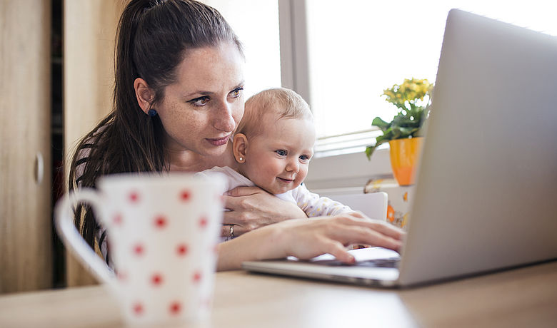 Eine junge Frau sitzt mit einem Baby auf dem Schoß vor einem Laptop.