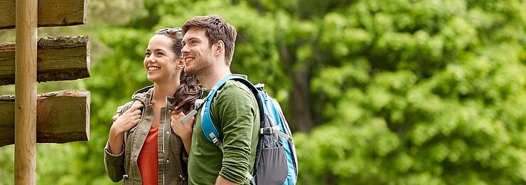 Zwei junge Menschen betrachten einen großen Wegweiser an einem Wanderweg im Grünen